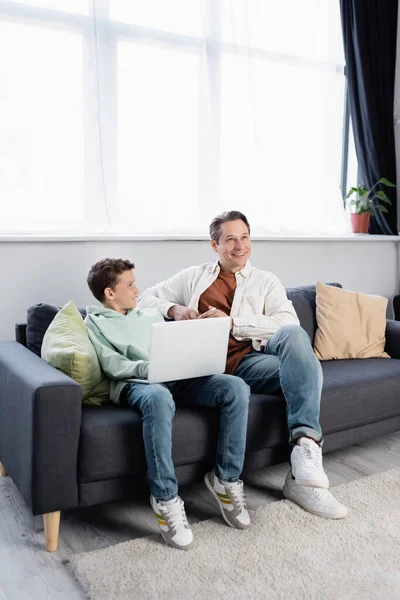 Positivo uomo tenendo smartphone vicino al figlio con computer portatile in soggiorno — Foto stock