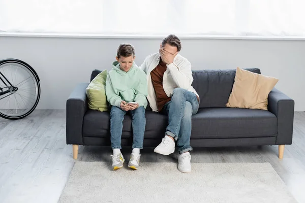 Man covering face while son using smartphone in living room — Stock Photo