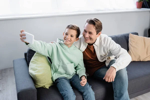 Усміхнений хлопчик бере селфі з батьком на дивані у вітальні — стокове фото