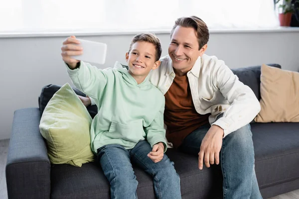 Sorrindo menino tomando selfie com o pai no sofá na sala de estar — Fotografia de Stock