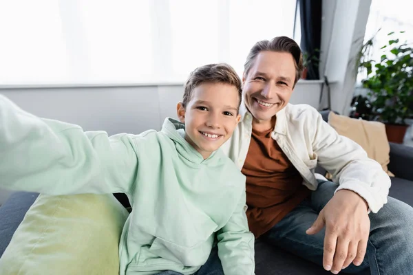 Cheerful boy and man looking at camera while sitting on couch at home — Stock Photo