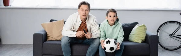 Preadolescente niño sosteniendo pelota de fútbol cerca de papá con mando a distancia en casa, pancarta - foto de stock