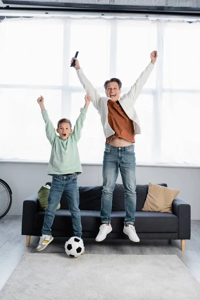 Família excitada com controle remoto pulando perto de bola de futebol em casa — Fotografia de Stock