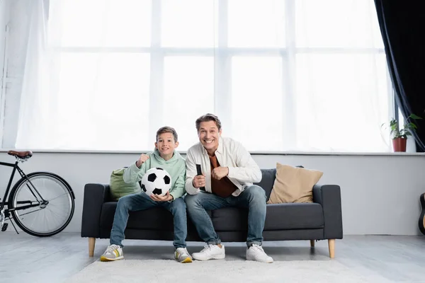 Father and son with remote controller and football ball watching match at home — Stock Photo