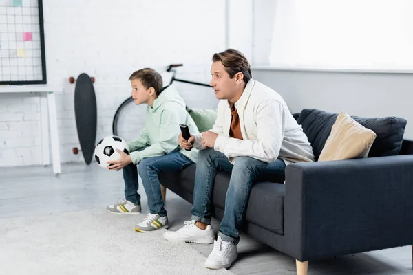 Konzentrierter Mann mit Fernbedienung beobachtet Fußballspiel in der Nähe seines Sohnes zu Hause — Stockfoto