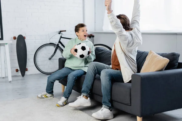 Emocionado niño preadolescente sosteniendo pelota de fútbol cerca de papá en el sofá en casa - foto de stock