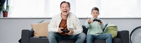 KYIV, UKRAINE - OCTOBER 11, 2021: Cheerful man playing video game with son on couch, banner — Stock Photo