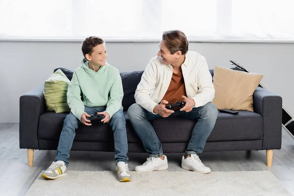 QUIIV, UCRÂNIA - OUTUBRO 11, 2021: Homem sorridente segurando joystick e olhando para o filho no sofá em casa — Fotografia de Stock