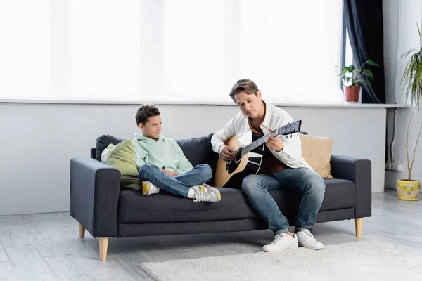 Mann spielt Akustikgitarre neben Sohn auf Couch zu Hause — Stockfoto