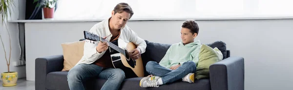 Отец играет на акустической гитаре рядом с сыном-подростком на диване, баннер — стоковое фото