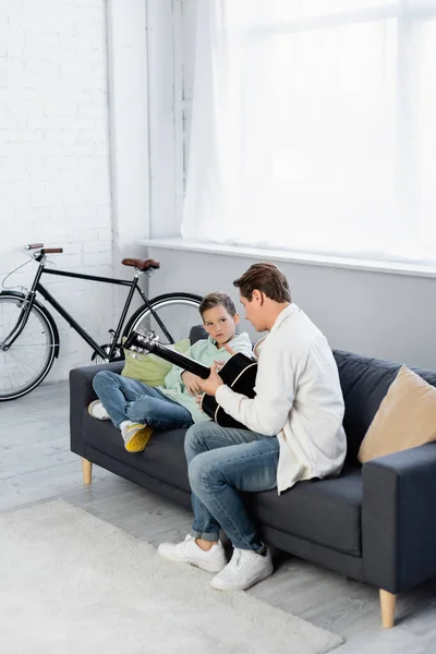 Hombre sosteniendo la guitarra acústica cerca de hijo preadolescente en el sofá en casa - foto de stock