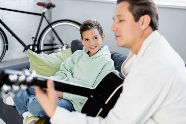 Lächelndes Preteen-Kind blickt verschwommenen Papa an, der zu Hause Akustikgitarre spielt — Stockfoto