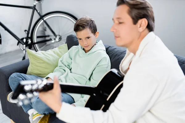 Niño mirando borrosa padre tocando la guitarra acústica en la sala de estar - foto de stock
