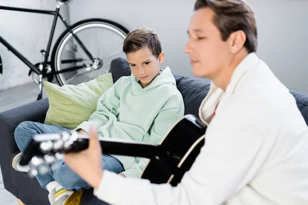 Junge sieht verschwommenen Vater, der Akustikgitarre im Wohnzimmer spielt — Stockfoto