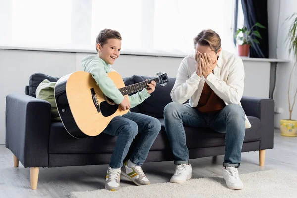 Улыбающийся мальчик играет на акустической гитаре рядом с лицом отца на диване — стоковое фото