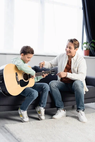 Позитивный отец указывает на акустическую гитару рядом с сыном дома — стоковое фото