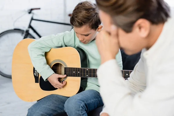 Garçon jouant de la guitare acoustique près flou père couvrant visage à la maison — Photo de stock