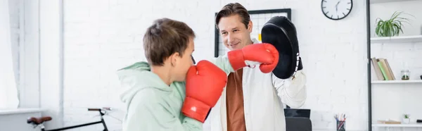 Ребенок-подросток в перчатках боксирует рядом с папой с блокнотом дома, баннер — стоковое фото