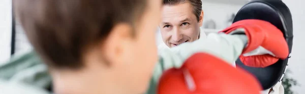 Père souriant dans le coussin de boxe travaillant avec son fils dans des gants à la maison, bannière — Photo de stock