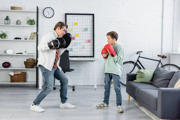 Side view of father in punching pads looking at son in boxing gloves at home — Stock Photo