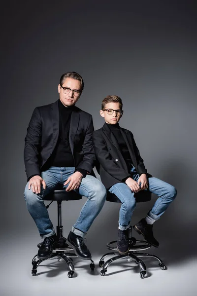 Stylish father and preteen son sitting on chairs on grey — Stock Photo