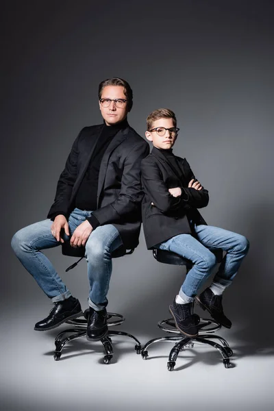 Stylish father and son posing on chairs on grey — Stock Photo