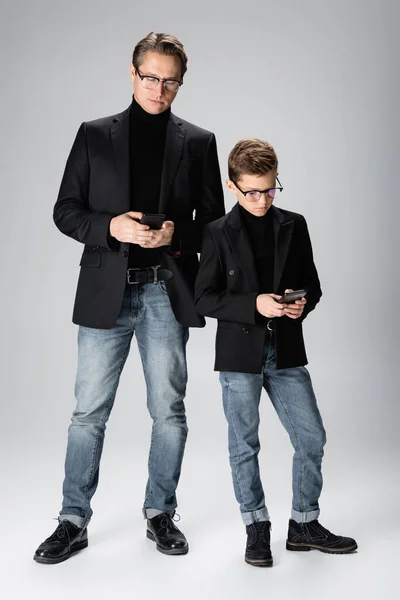 Longitud completa del hombre y el hijo preadolescente utilizando teléfonos inteligentes sobre fondo gris - foto de stock