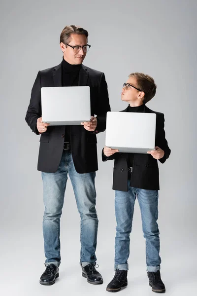 Niño con chaqueta sosteniendo portátil y mirando a papá sobre fondo gris - foto de stock