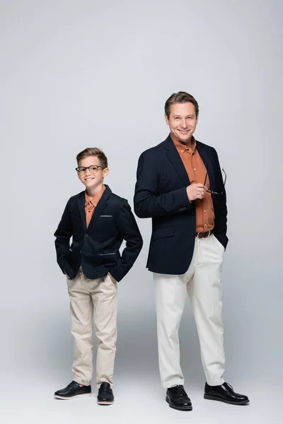 Elegante hombre sosteniendo anteojos cerca de sonriente hijo sobre fondo gris - foto de stock