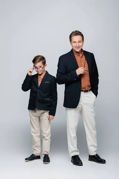 Longitud completa de los padres de moda que sostienen las gafas cerca de hijo sobre fondo gris - foto de stock