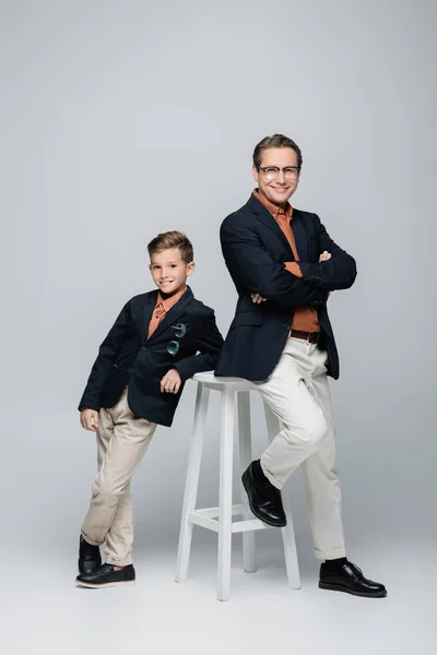 Padre e hijo positivos y elegantes posando cerca de la silla sobre fondo gris - foto de stock