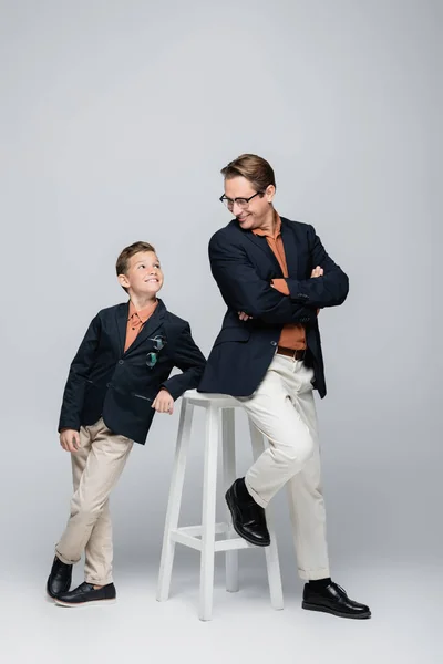 Hombre positivo y niño mirándose el uno al otro cerca de la silla sobre fondo gris - foto de stock