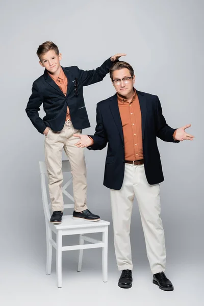 Son in jacket standing on chair near dad on grey background — Stock Photo