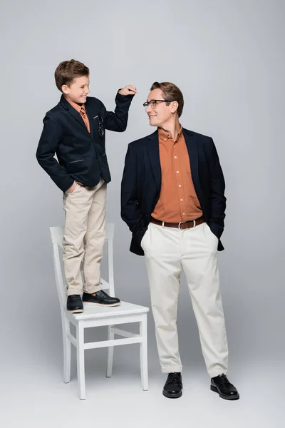 Smiling boy pointing at stylish dad while standing on chair on grey background — Stock Photo