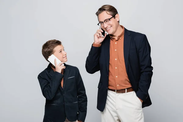 Sonriente padre e hijo en chaquetas hablando en teléfonos inteligentes aislados en gris - foto de stock