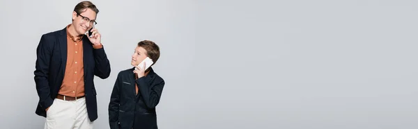 Sonriente hombre de chaqueta hablando en el teléfono inteligente cerca de hijo aislado en gris, bandera - foto de stock