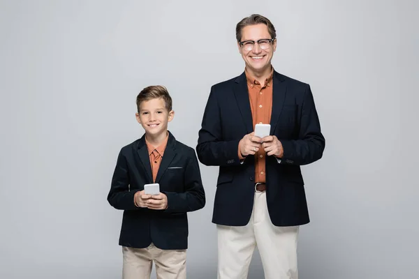 Sonriente padre e hijo mirando a la cámara mientras sostiene los teléfonos inteligentes aislados en gris - foto de stock