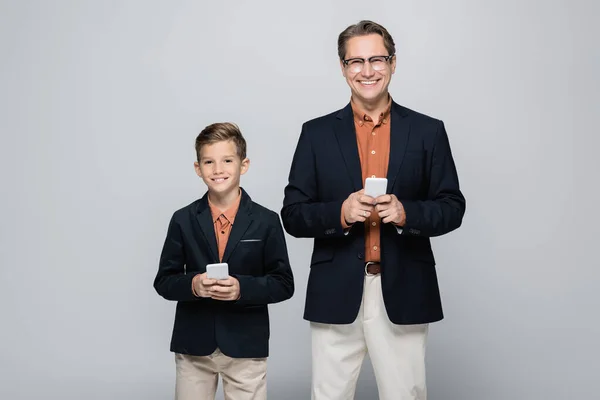 Smiling parent and son looking at camera while holding smartphones isolated on grey — Stock Photo