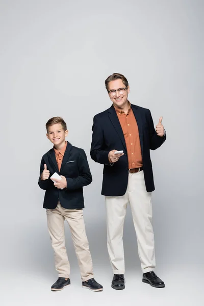 Sonriente padre e hijo sosteniendo teléfonos inteligentes y mostrando como en un fondo gris - foto de stock