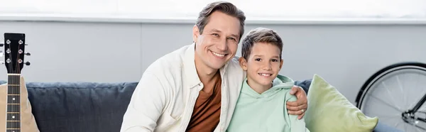 Hombre positivo abrazando chico en el sofá en la sala de estar, pancarta - foto de stock