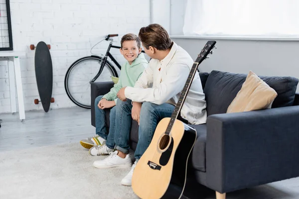 Garçon souriant parlant à son père près de la guitare acoustique à la maison — Photo de stock