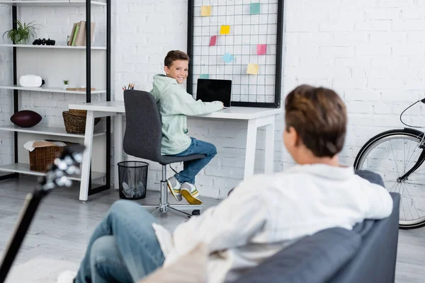 KIEW, UKRAINE - 11. OKTOBER 2021: Lächelnder Junge mit Laptop neben verschwommenem Papa auf der heimischen Couch — Stockfoto