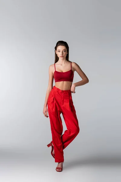 Повна довжина молодої моделі в червоному вбранні позує з рукою на стегні на сірому — стокове фото