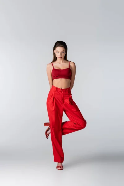 Full length of pretty model in red outfit posing while standing on one leg on grey — Stock Photo