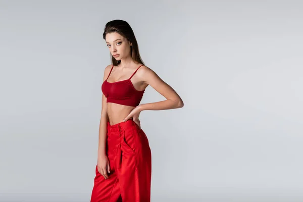 Молодая модель в красном наряде, позирующая с рукой на бедре, изолированная на сером — стоковое фото