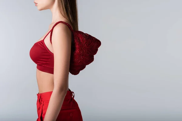 Обрезанный вид молодой женщины в красном наряде со стильной сумкой на плече, изолированной на сером — стоковое фото