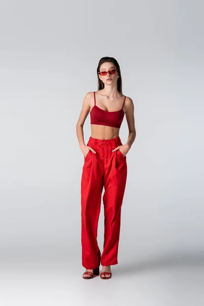 Junges Model in Sonnenbrille und rotem Outfit posiert mit Händen in Taschen auf grau — Stockfoto