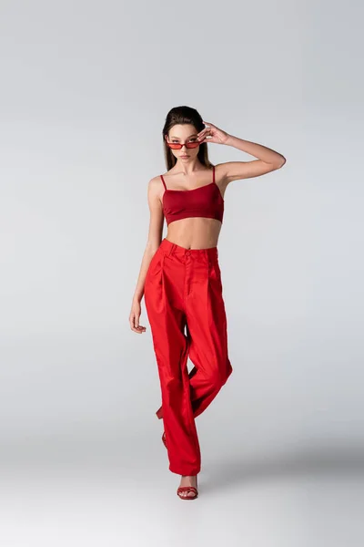 Full length of young model in red outfit adjusting sunglasses on grey — Stock Photo