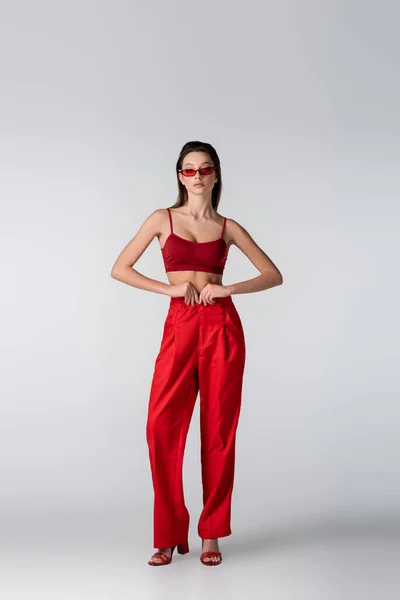 Comprimento total da jovem mulher em óculos de sol e roupa vermelha posando em cinza — Fotografia de Stock