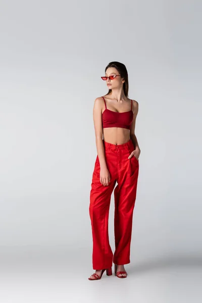 Полная длина молодой модели в солнцезащитных очках и красной одежде, позирующей с рукой в кармане на сером — стоковое фото